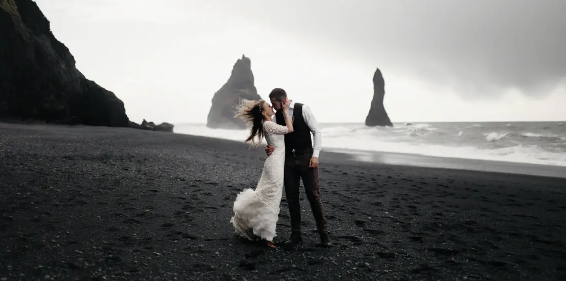 A bride and groom holding each other on a black sand beach, sea stacks and surf behind them