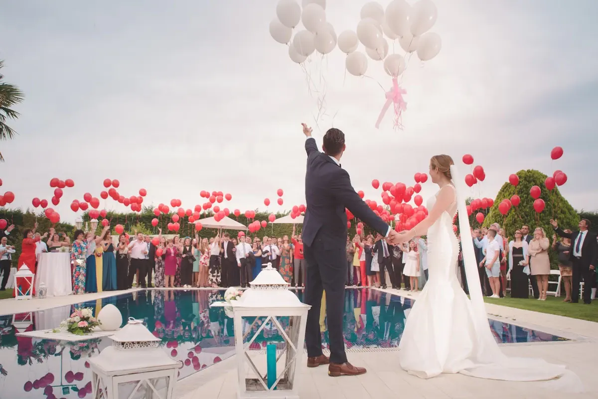A bride and groom release white balloons at a poolside wedding while their guests hold red balloons