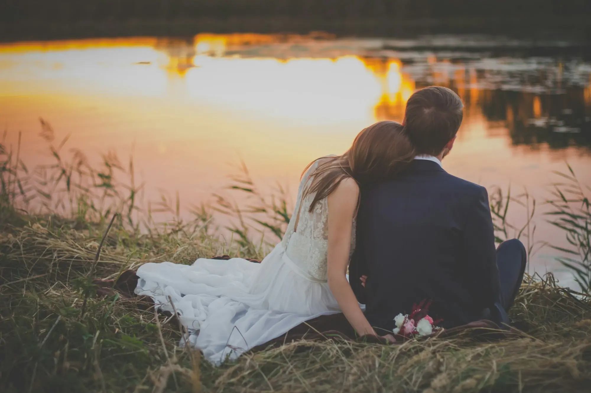 A bride and groom sitting together by a lake at sunset, her bouquet on the blanket beside them