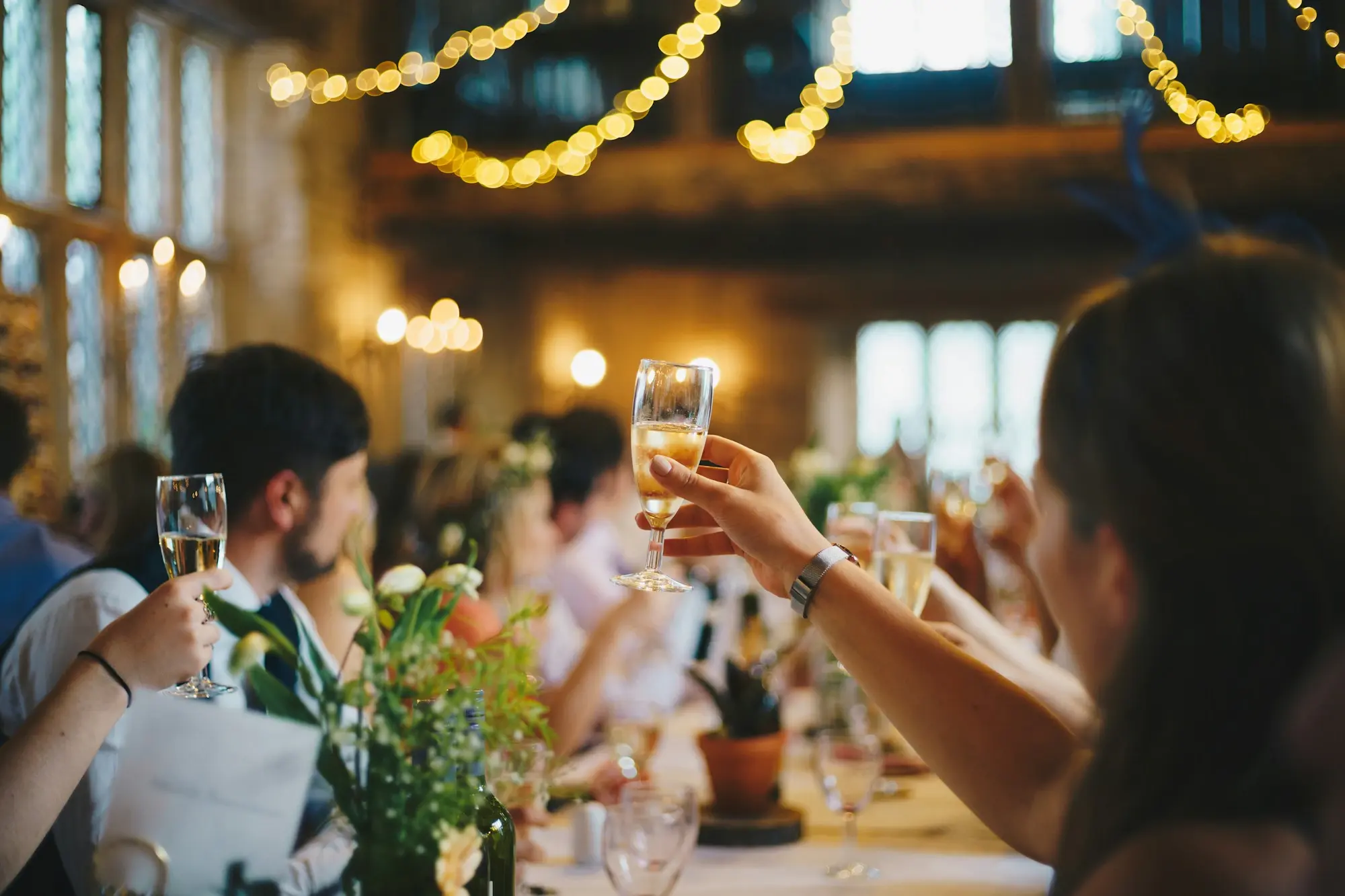 Guests raising champagne glasses at a long reception table under strings of warm lights