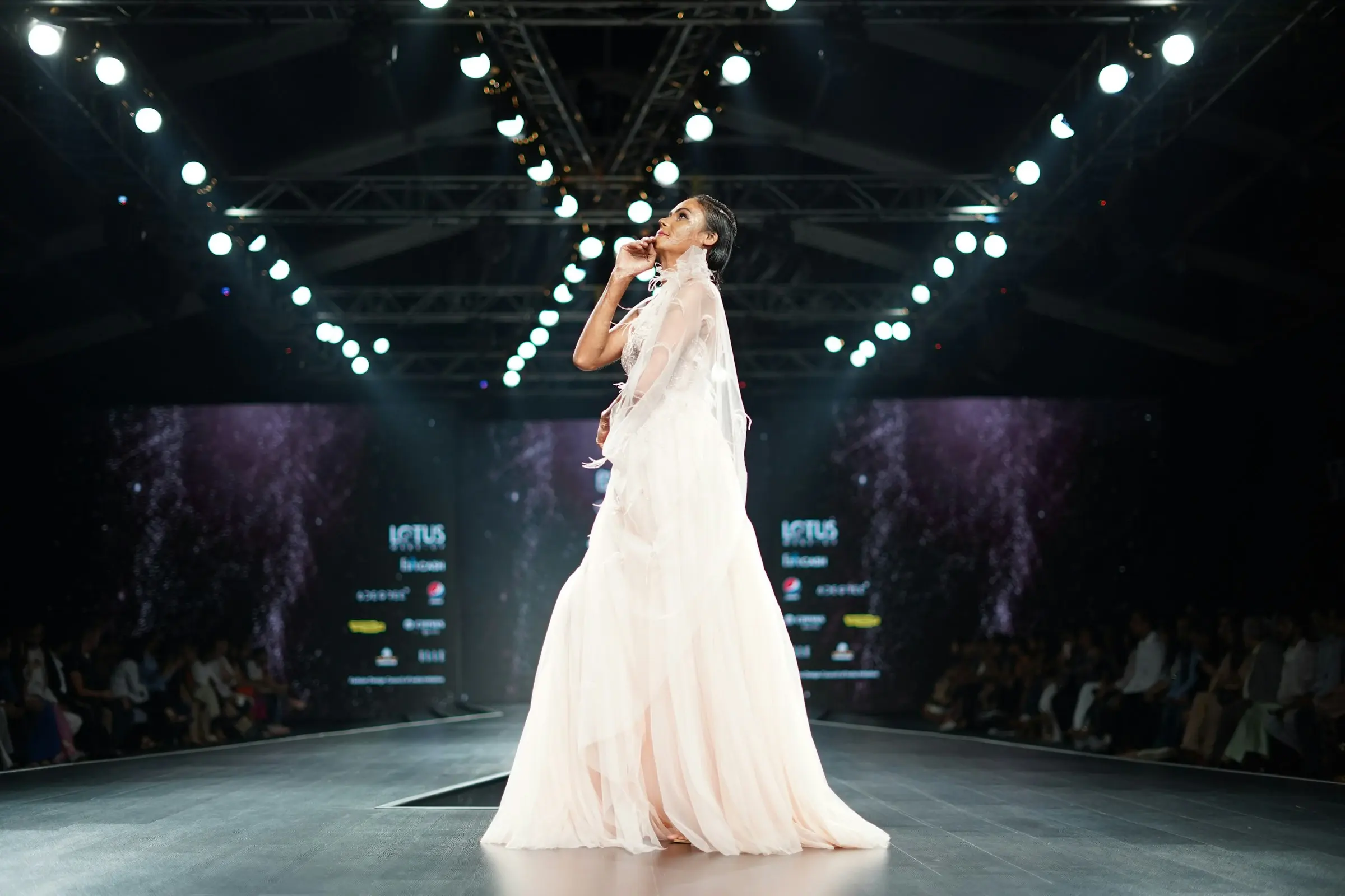 A model in a blush gown walking a lit runway under stage lighting