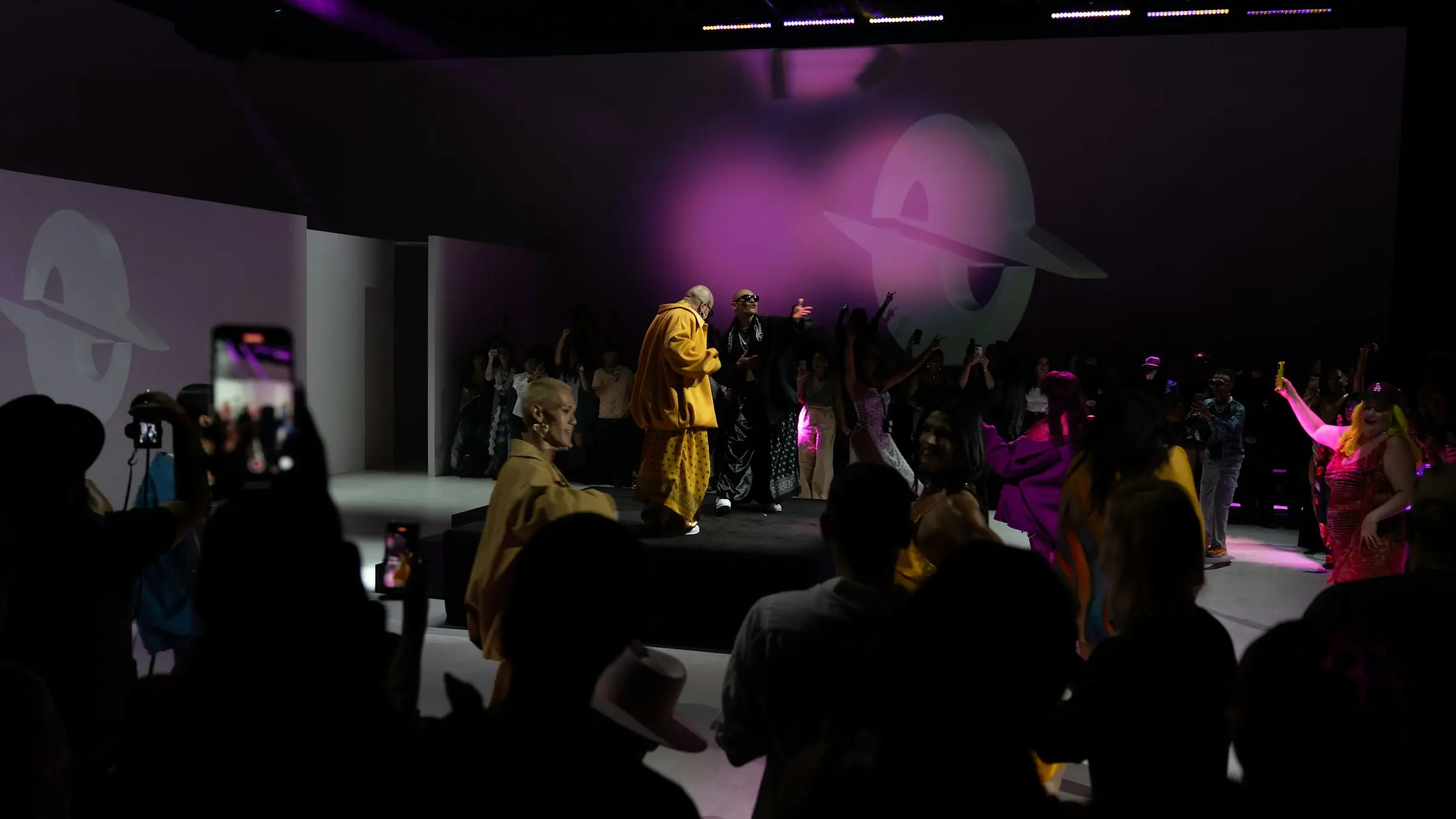 A fashion show under magenta stage lighting, the audience silhouetted in the foreground